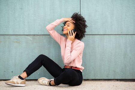 Portrait happy african american woman sitting on floor outside talking with mobile phone
