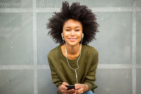 Portrait smiling young african american woman leaning against wall with smart phone and earphoneの写真素材