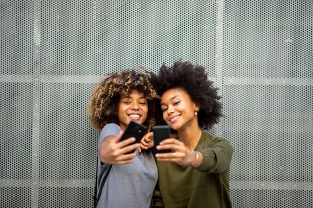Portrait two happy young black women taking selfie with mobile phonesの写真素材