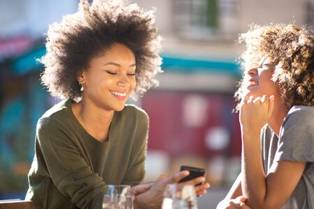 Portrait two happy young african american women sitting at outdoor cafe looking at cellphoneの写真素材
