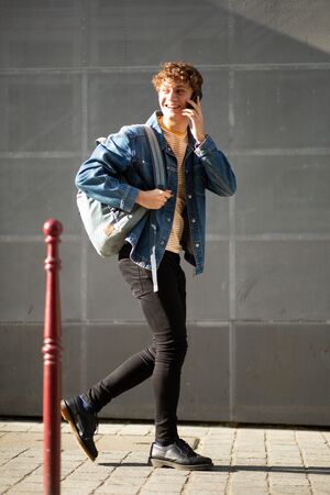 Full length side portrait young man walking and talking with cellphone on sidewalk