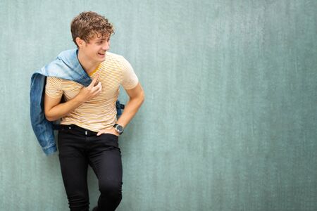 Portrait laughing young man posing by green wall with denim jacket over shoulder