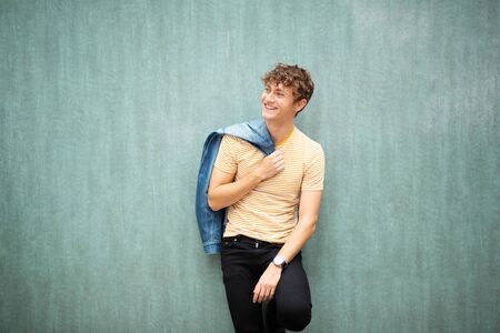 Portrait smiling young man posing by green wall with denim jacket over shoulder
