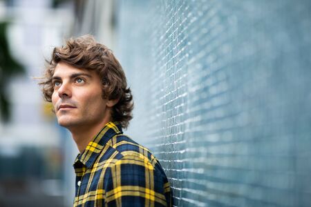 Close up portrait handsome young man leaning against wall thinking looking upの写真素材