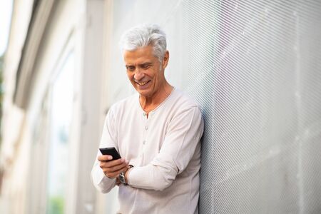 Portrait smiling older man leaning against wall looking at mobile phone