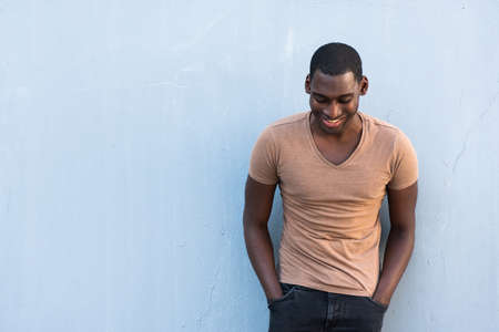 Portrait happy young African American guy leaning against gray wall with hands in pocket and looking downの写真素材