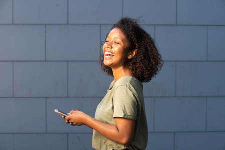 Profile portrait happy young woman holding cellphone and looking away by gray wallの写真素材