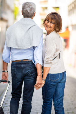 Portrait from back of older couple walking on street with suitcase. Woman smilingの写真素材