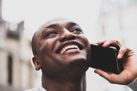 Portrait African american man using mobile phone while standing next to white wall with bag and headphonesの写真素材