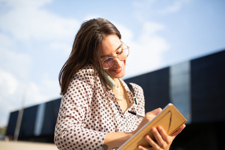 Portrait of young businesswoman student standing outside talking on mobile phone and writing in bookの写真素材