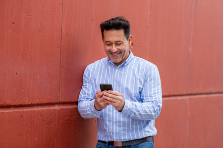 Portrait of mature man leaning to a red wall and using mobile phoneの写真素材