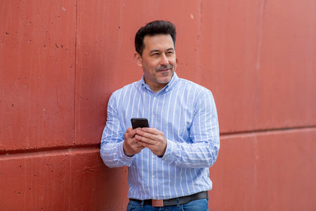 Portrait of mature man leaning to a red wall with mobile phone looking awayの写真素材