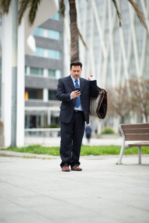Full length portrait of mature businessman standing outdoors in the city with suit travel bag and using his cell phoneの写真素材