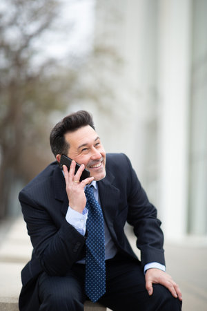 Portrait of happy mature businessman sitting outside talking on cellphoneの写真素材