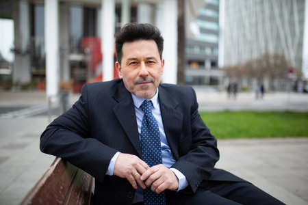 Portrait of mature latin businessman sitting on bench outside in the cityの写真素材