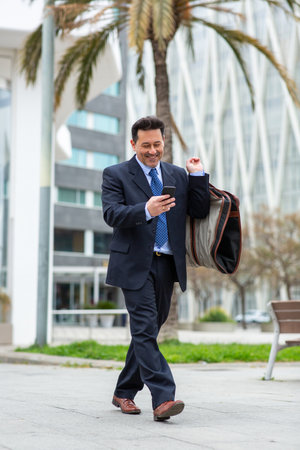 Full length portrait of happy mature businessman walking in the city with suit travel bag and using his mobile phoneの写真素材