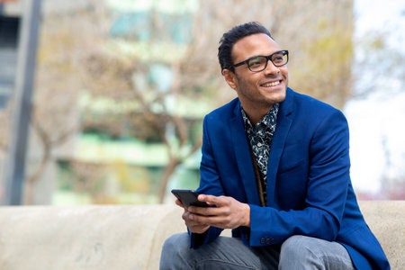 Portrait of happy young african american businessman sitting outside with cell phone and looking awayの写真素材