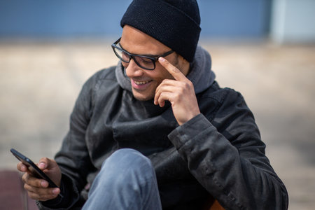 Portrait of smiling black man in winter jacket and cap sitting on outside bench in city using mobile phoneの写真素材