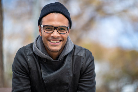 Close up portrait of handsome young african man in warm jacket and knitted hat looking at camera and smiling outside in the cityの写真素材