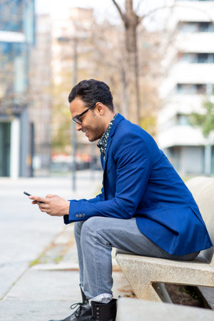 Side portrait of mixed race man in suit sitting outside on bench and texting on mobile phoneの写真素材