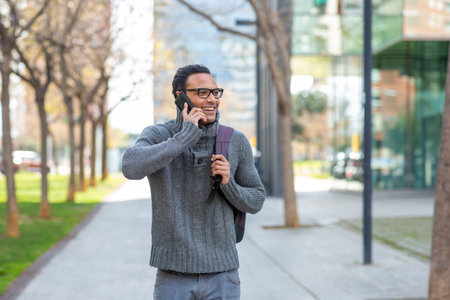 Portrait of cheerful young african man with bag walking outside and talking on phoneの写真素材