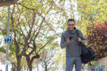 Portrait of happy young african american man texting on mobile phone carrying bag outsideの写真素材