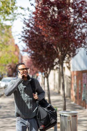 Portrait of happy young african man talking on mobile phone carrying bag outsideの写真素材