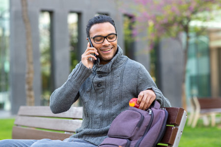 Portrait of happy young black man talking on cell phone while sitting on bench with bag outside in cityの写真素材