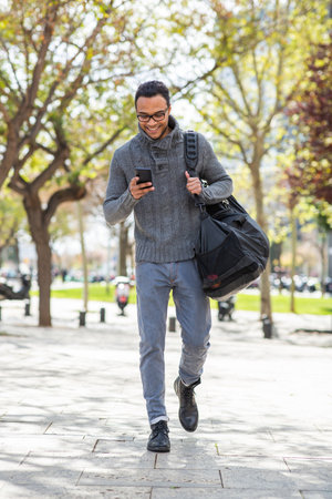 Full body portrait of cool young guy using cell phone carrying bag outsideの写真素材