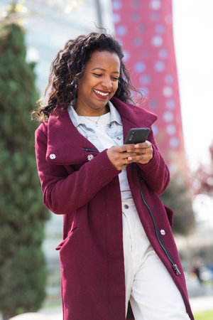 Portrait of beautiful young black woman in winter coat walking outside and using cell phoneの写真素材