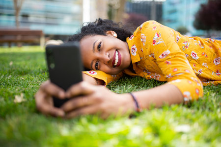 Portrait of happy young black woman lying on grass and taking selfie using mobile phone outside