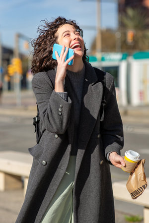 Portrait of smiling young woman talking on phone while walking in a city street with takeaway coffee in handの写真素材