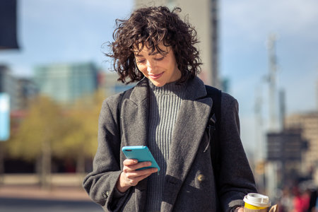 Portrait of young woman in coat using mobile phone while walking outdoors in cityの写真素材