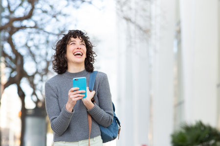 Portrait of beautiful young woman with bag and phone walking outside in city looking away and laughingの写真素材