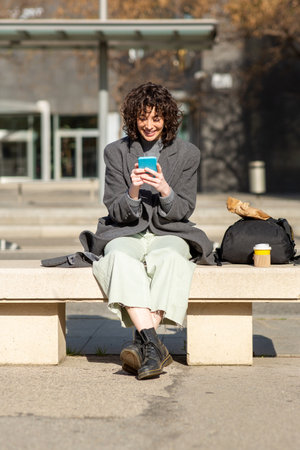 Portrait of happy young woman commuter messaging using mobile phone while sitting on bench outside with bagの写真素材