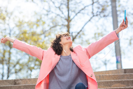 Portrait of attractive woman wearing earphones sitting city steps with her arms outstretched holding mobile phone and smilingの写真素材