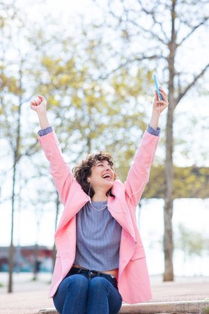 Portrait of cheerful young woman sitting outside with her arms outstretched holding mobile phoneの写真素材