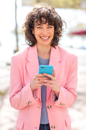 Portrait of pretty young woman with phone standing outside in city looking at camera and smilingの写真素材