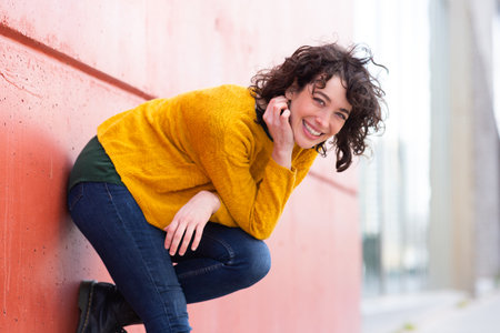 Portrait of attractive young woman bending over looking at camera and smiling by the wall outdoorsの写真素材