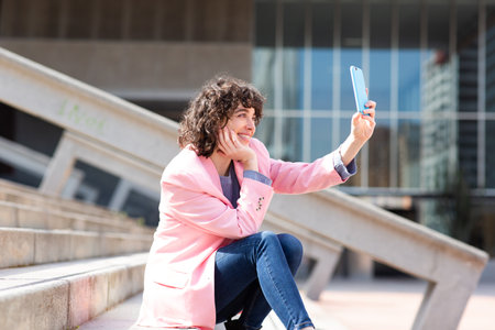 Side view of young woman taking selfie with her mobile phone sitting on steps outside in the cityの写真素材