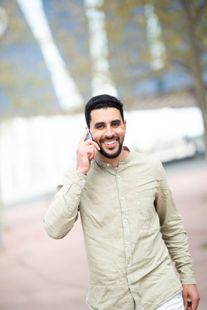 Portrait of cheerful young arab man talking on cellphone walking outside in the cityの写真素材