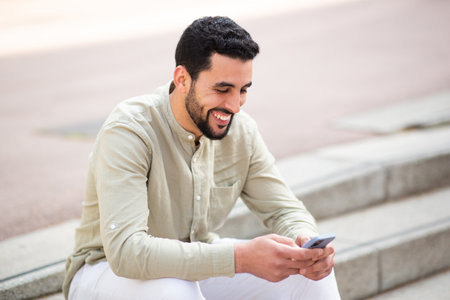Portrait of cheerful young middle eastern man sitting outside on steps and using cell phoneの写真素材
