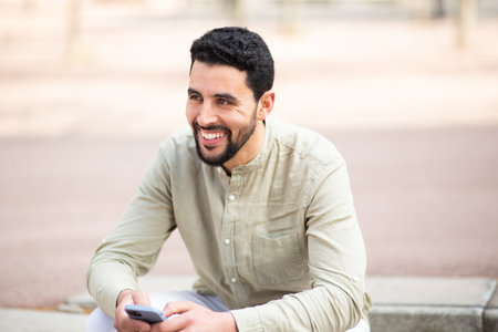 Portrait of handsome young arab man sitting outside with mobile phone looking away and smilingの写真素材