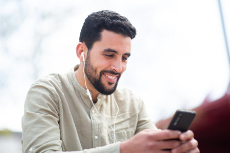 Portrait of smiling north african guy sitting outside with earphones listening to music from mobile phoneの写真素材