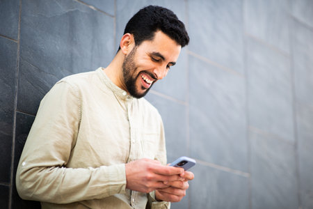 Portrait of smiling young north african man using mobile phone leaning to a wallの写真素材