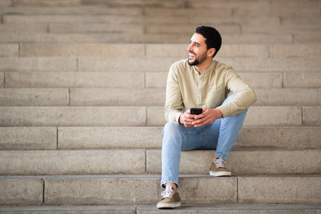 Full length portrait of young arab man sitting on steps with mobile phone looking away and smilingの写真素材