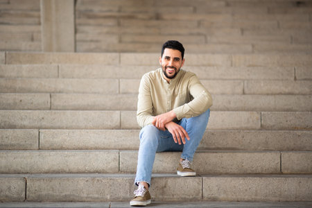 Full length portrait of handsome young arab man sitting on steps looking at camera and smilingの写真素材