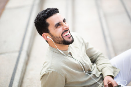 Side portrait of smiling arabic man listening to music with headphones sitting outdoorsの写真素材