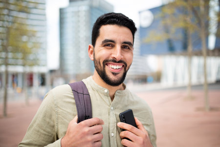 Portrait of smiling young middle eastern man holding mobile phone walking outside in the cityの写真素材