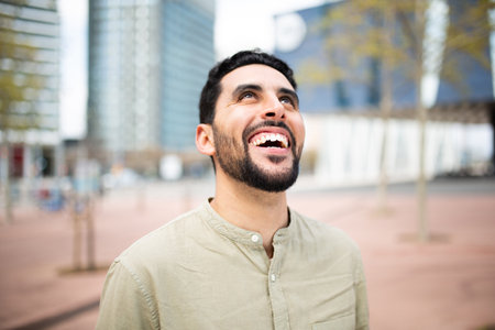 Close up portrait of carefree young arabic man standing outside in the city looking up and laughingの写真素材
